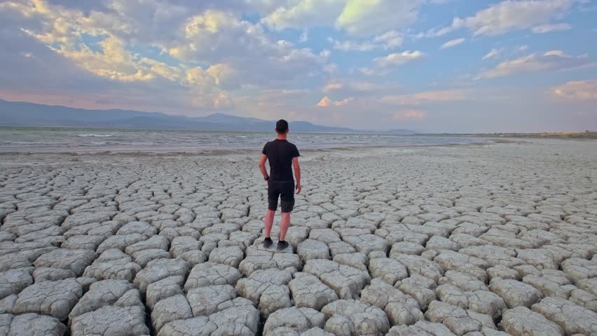 A man stands on cracked, dry land, gazing at the horizon. The dried-up lakebed and dramatic sky highlight the impacts of climate change.