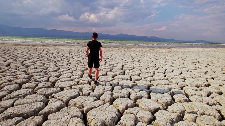 
A man walks on cracked, dry land, gazing at the horizon. The dried-up lakebed and dramatic sky highlight the impacts of climate change.