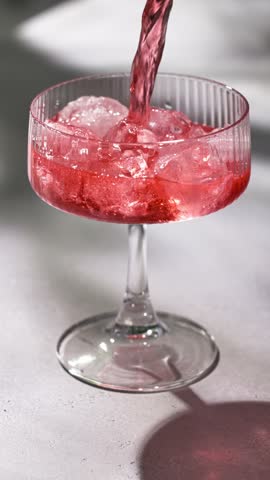 fresh red drink pouring into a cocktail glass with ice cubes on a gray sunny restaurant tabletop, slow motion