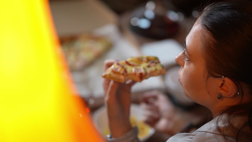 A young adult Caucasian lady is eating slice of pizza sitting at home in the evening and watching TV. Closeup slow motion footage of a woman spending weekend alone with fast food for snack and movie