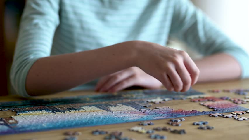 Close-up view of young woman hands assembling puzzles at home. Connecting jigsaw puzzle pieces, assembling a jigsaw puzzle. Fun family leisure. Table games, home relaxation. Slow motion footage.