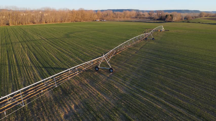Diagonal aerial filming of an agricultural irrigation pivot at sunset, located in a developing crop field.