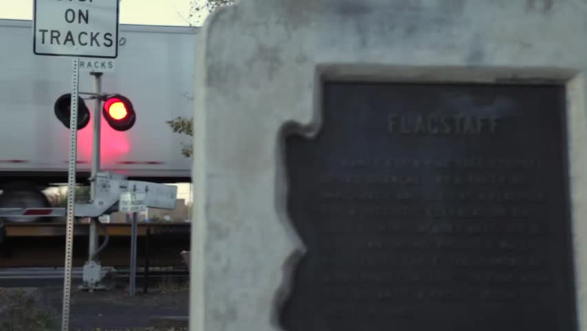 Close up of Flagstaff Arizona Historical Marker with railroad lights flashing and train passing by behind it.