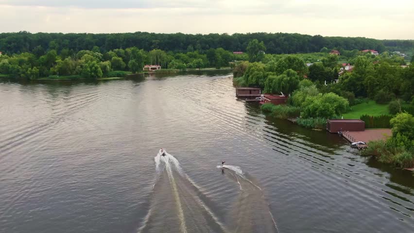 A dynamic drone video capturing boats towing wakeboarders on a serene Romanian lake in the countryside. The lush green hills and picturesque rural landscape frame the action.