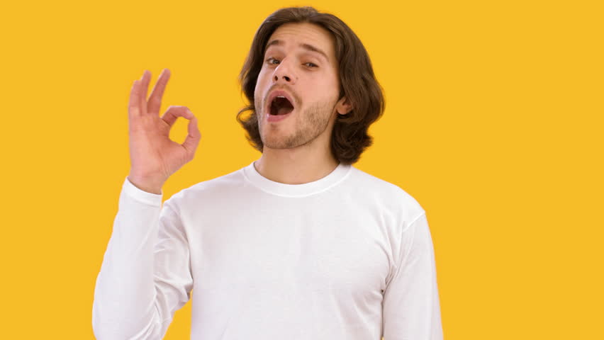 A smiling young man is making the OK sign with his right hand while wearing a cozy yellow sweater. The vibrant yellow background enhances the joyful atmosphere, reflecting positivity and friendliness.