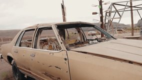 A wrecked, old, beige car sits abandoned in a desert landscape. Its windows are broken, the seats torn, and paint chipped. A makeshift sign made of bottles hangs nearby, - Powered by Shutterstock - Get 15% off with code: PIKWIZARD15