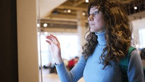 Young woman with curly hair and backpack interacting with large touchscreen display in contemporary public space, exploring information with intuitive gestures - Powered by Shutterstock - Get 15% off with code: PIKWIZARD15