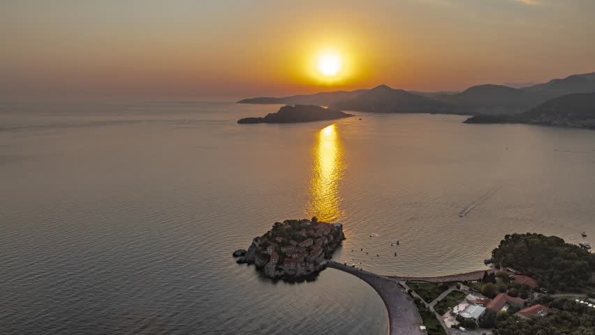 Evening drone hyperlapse over the island of Sveti Stefan from above, with the sunset lit pathway in the sea and mountains in the background, Montenegro, Europe