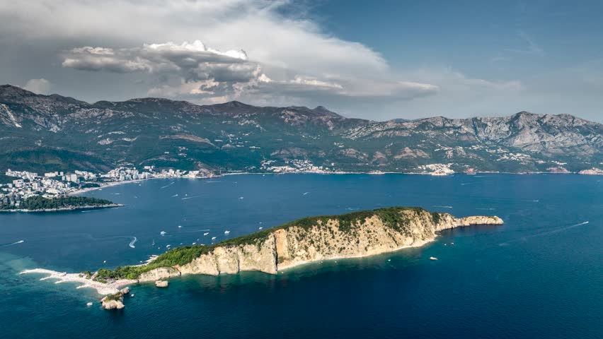 Epic hyperlapse drone shot over Sveti Nikola Island, the local Hawaii, with numerous boats in the sea, and Budva with mountains in the background, featuring epic, fast-moving cumulus clouds