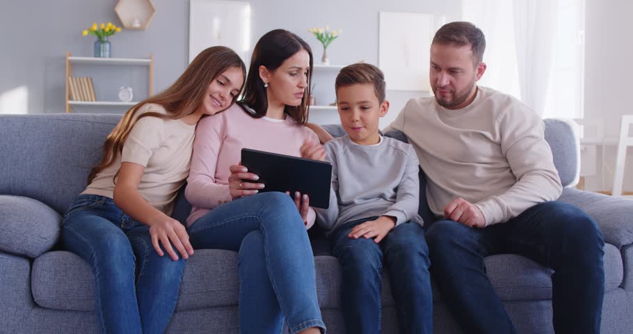 Happy young family with two little kids at home, relaxing on sofa in living room using tablet online, talking speak on video webcam call on computer, parents with teen children at using gadget