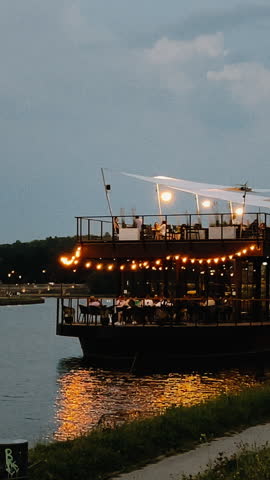 A beautifully picturesque dining boat shines brightly against a serene and tranquil waterfront during twilight hours. vertical cinematic shot, film look