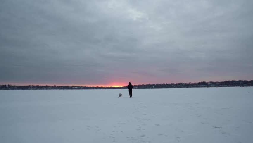 Man wearing headphones walks with his pug across a vast frozen lake in Minnesota during dusk