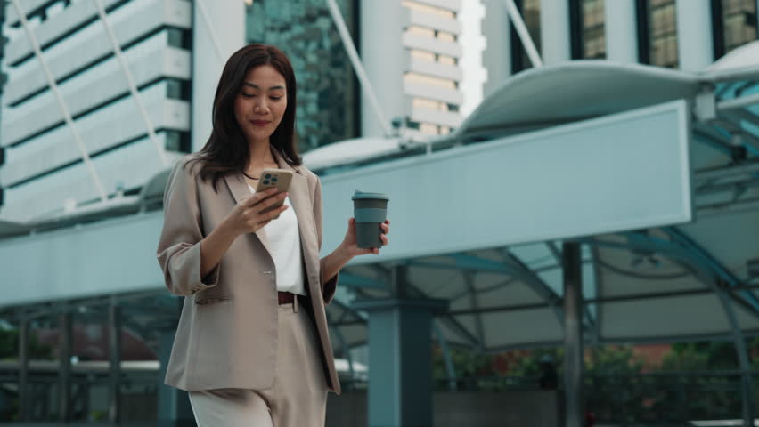 Portrait of happy Thai businesswoman with smartphone and coffee cup walking along street. Young woman wearing smart casual pantsuit uses chat on mobile phone. Successful career in business
