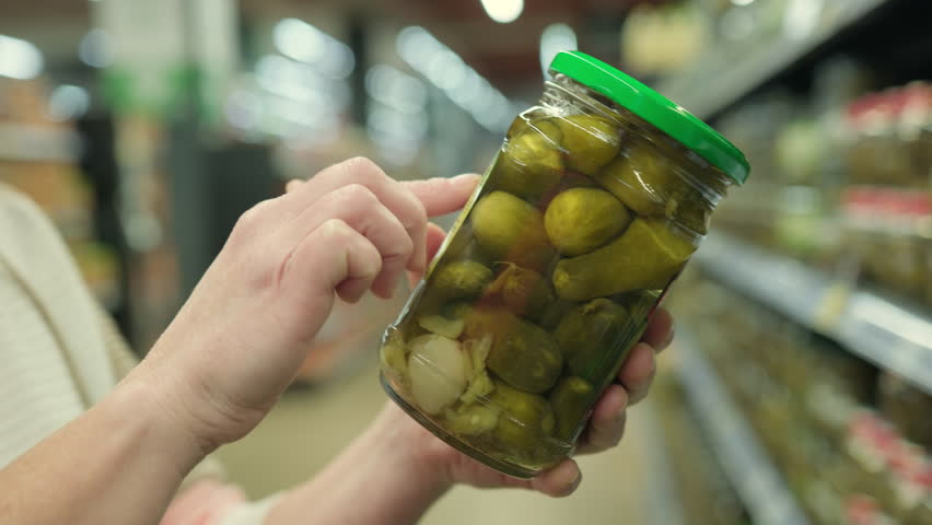 Woman hand reading label on glass jar with pickles before buying in supermarket close-up.