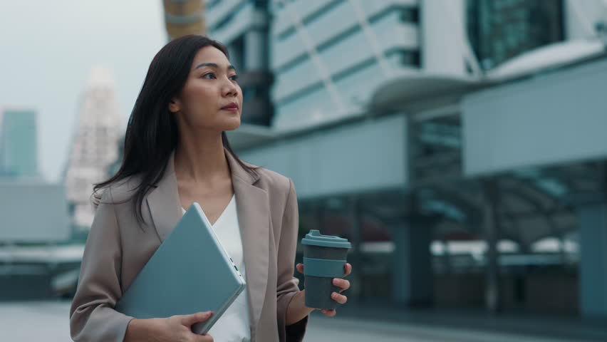 Portrait of young Asian woman with laptop and coffee cup walking along street. Thoughtful Thai business lady holds computer and drink near office center. Successful commercial career