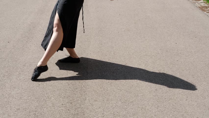 Woman is dancing outside in a summer park. Close view of legs. Female dancer in a black dress makes graceful movements outdoor. 4k horizontal slow motion footage.