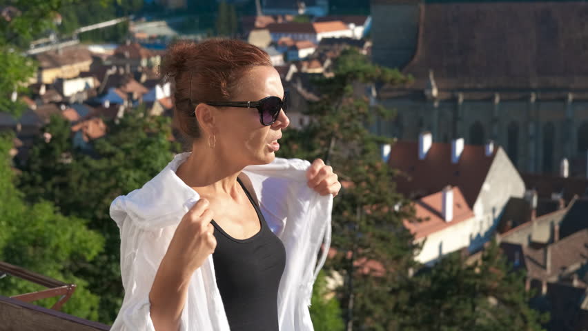 Tourist woman suffering from summer heat. Redhead tourist woman wearing sunglasses and white shirt suffering from summer heat, with the medieval city in the background