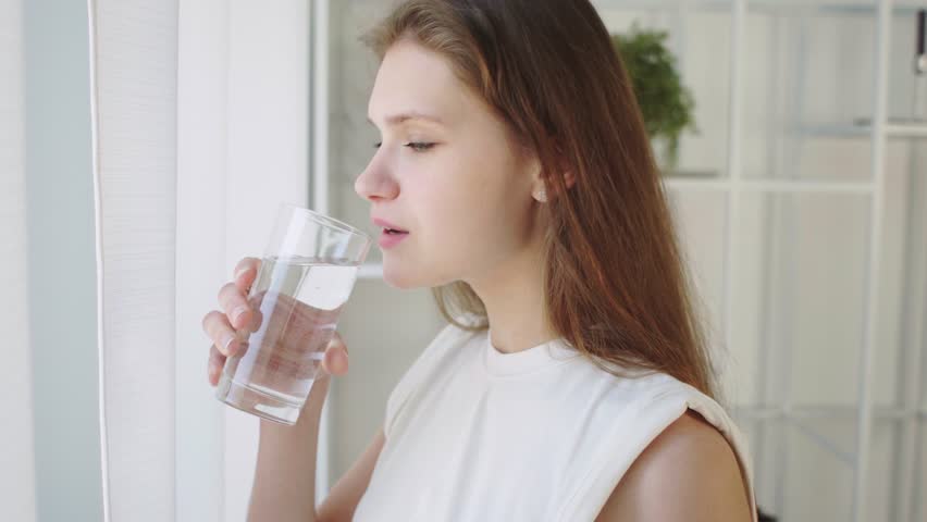 Young beauty woman taking vitamin Fish Oil Omega-3 capsule by drinking a glass of water standing by window at home, Health, people, food, sports, lifestyle and beauty concept Side View 4K