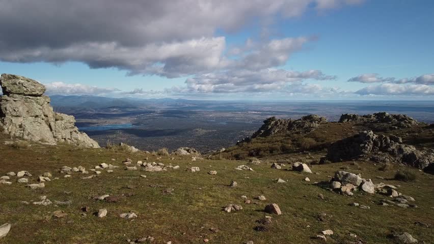 A panoramic view from a mountaintop reveals a breathtaking landscape. Rolling hills, a lake, and distant mountains stretch out towards the horizon. Dramatic clouds add depth and texture to the sky.