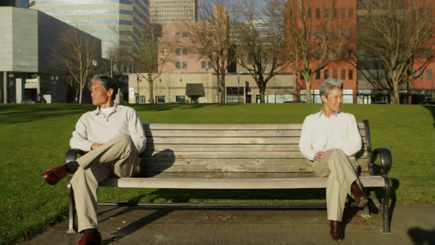 Older Asian-American man and woman sit at opposite sides of park bench,
