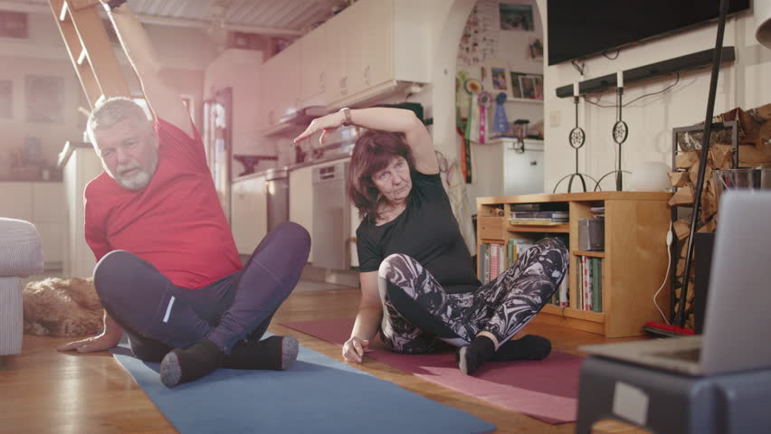 Seated retired older couple follow guided yoga exercises from laptop