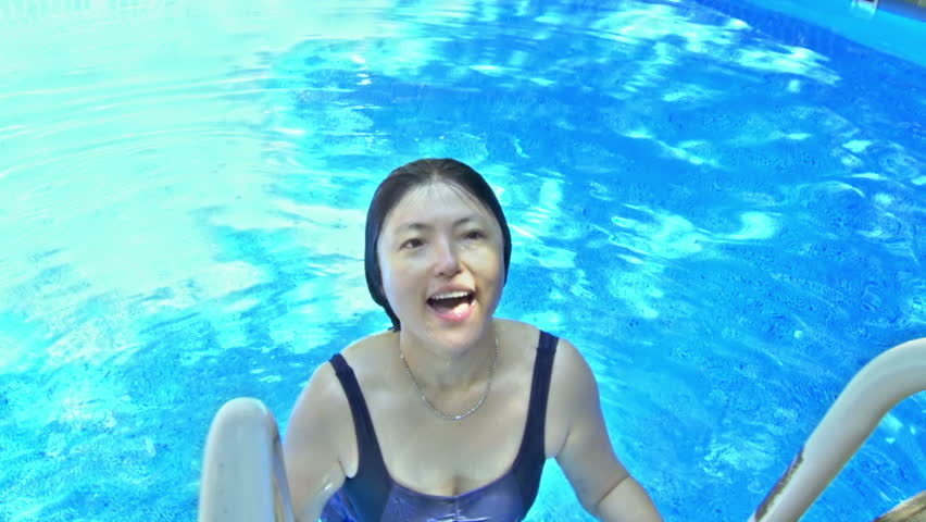 Asian woman swims up out of pool, is wrapped in a towel then shuffles away