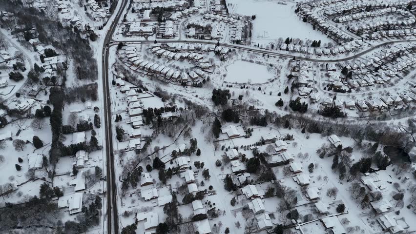 An aerial footage of roads with calm traffic through a snow-covered urban neighborhood with white roofs during winter in Waterloo, Ontario, Canada