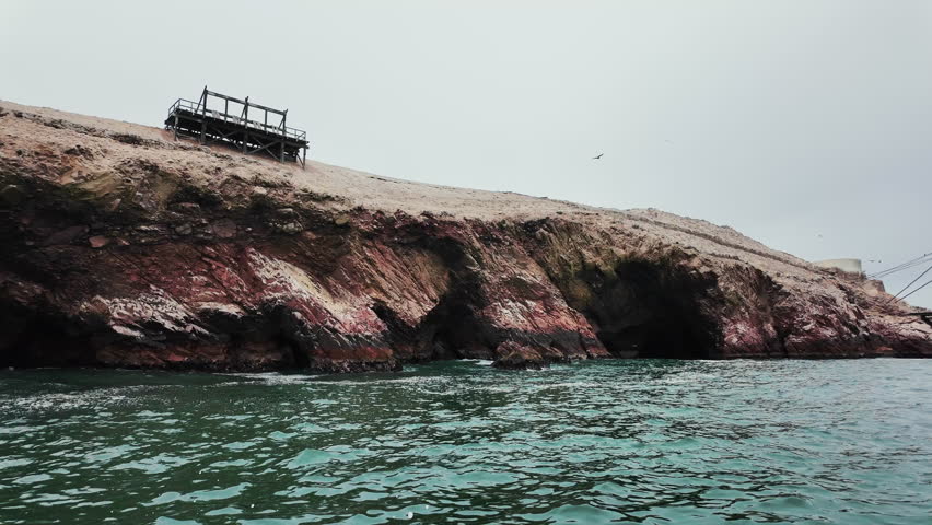The wild charm of the Ballestas Islands unfolds with cloudy skies, dramatic cliffs, and birds soaring over the Pacific waters.
