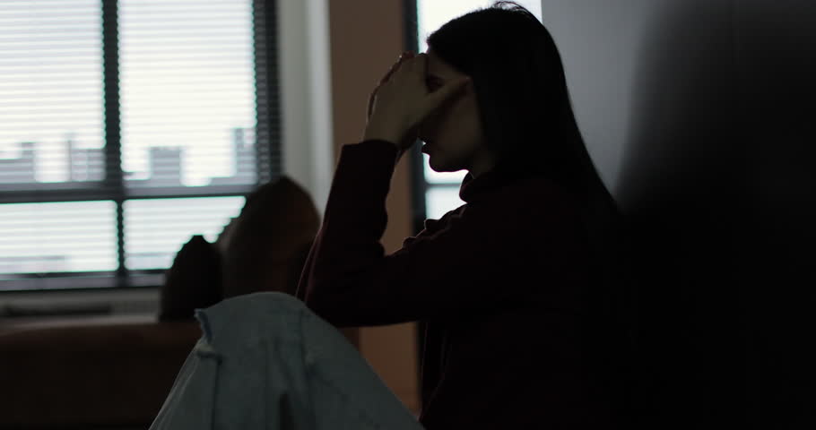Silhouette of a sad woman sitting in the dark leaning against the wall in home. Domestic violence, family problems, stress, violence.