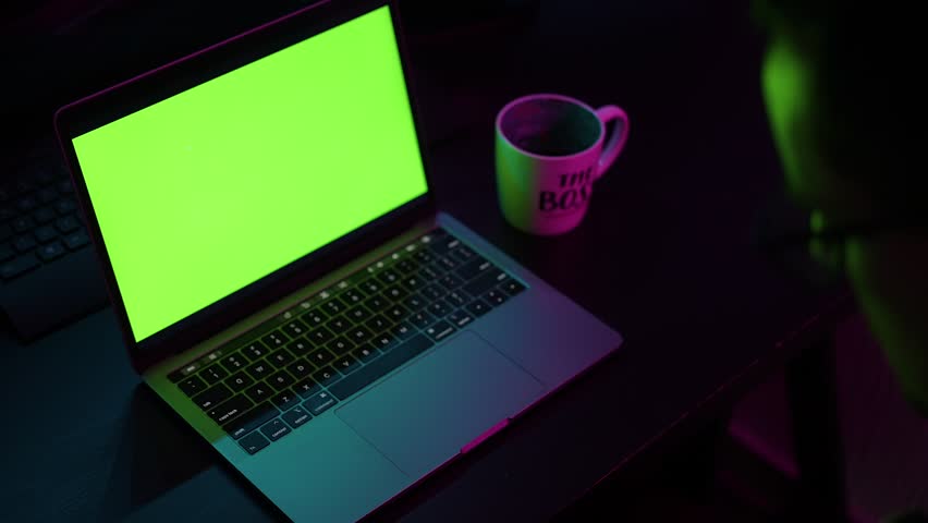 A person types on a laptop in a dimly lit room. The screen glows bright green, suggesting a placeholder or chroma key. A mug labeled "The Boss" and a phone add to the workspace atmosphere. - Powered by Shutterstock - Get 15% off with code: PIKWIZARD15
