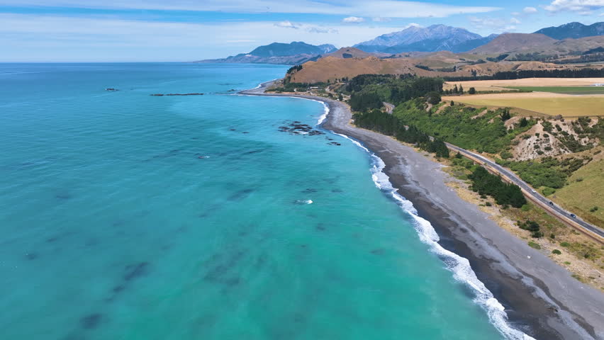 Aerial flies south above the aqua Pacific Kaikoura Coast, following Black Beach towards the settlement of Kekerengu, on a hot fine day with vast mountains behind New Zealand Aotearoa