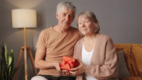 A senior couple sits together in a warmly decorated living room, smiling as they exchange a wrapped gift. The atmosphere is filled with joy and intimacy as they share this moment - Powered by Shutterstock - Get 15% off with code: PIKWIZARD15