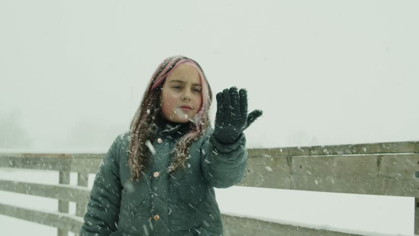 A Nine-Year-Old Girl, Dressed In Winter Clothes, Stands By A Snowy Lake As Snow Falls On Her Hand. She Gently Looks Up At The Sky, Enjoying The Quiet, Snow-Covered Landscape.