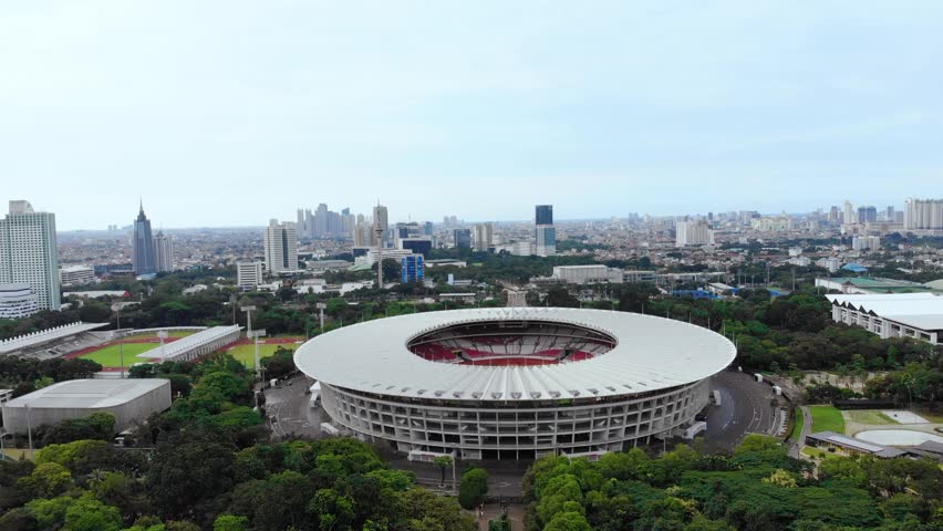 Aerial drone view of Gelora Bung Karno football stadium in Senayan, Jakarta. A lush sports center with lots of trees and views of skyscrapers.