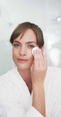 Vertical video: Caucasian woman in white robe applies lip balm at home. Freckles dot her fair skin, brown hair up, blue eyes forward, minimal makeup, simple earring