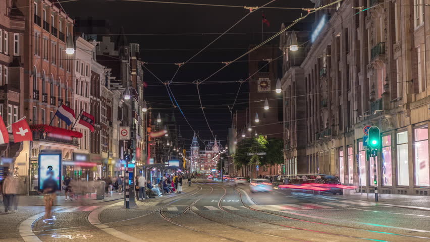 Electric tram on Damrak Street heading from Dam Square to Centraal Station in a timelapse at night. Traffic moves on the road while people wait at the tram stop. Amsterdam, Netherlands