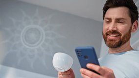 Happy man sipping coffee watching mobile phone content at light kitchen closeup. Positive guy reading smartphone message taking cappuccino sip. Morning businessman scrolling social media at flat  - Powered by Shutterstock - Get 15% off with code: PIKWIZARD15