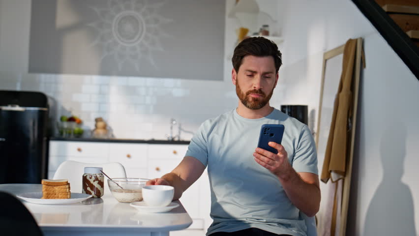 Relaxed brunette looking mobile phone sitting with coffee cup alone closeup. Morning guy watching smartphone screen reading message at kitchen. Cheerful man scrolling social media cell drinking tea 