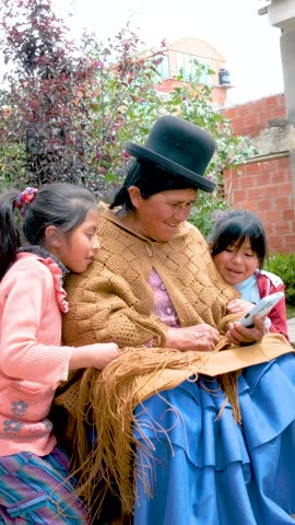 vertical aymara cholita grandmother in pollera looking at her cell phone with her two granddaughters in the backyard of her house in the city of la paz bolivia - communication concept