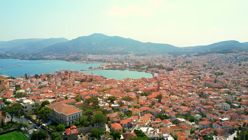 Drone shot revealing the city and port of Mytilene, Greece, with a football pitch, traffic, terracotta roofs, and hills in sight.