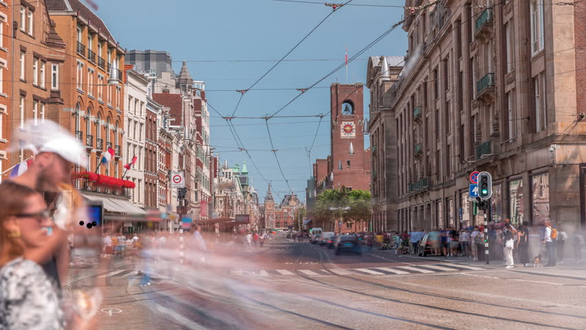 Electric tram on Damrak Street heading from Dam Square to Centraal Station in a timelapse. Traffic moves on the road while people wait at the tram stop. Amsterdam, Netherlands