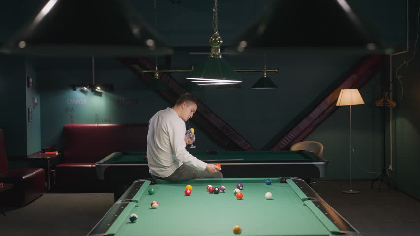 Billiard trainee wearing white shirt and checkered trousers sits on green pool table, holding a glass of lemon drink with black straw while picking a billiard ball. Relaxed training atmosphere