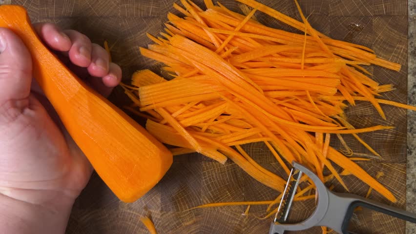 Woman peeling carrots with peeler against wooden cutting board.
