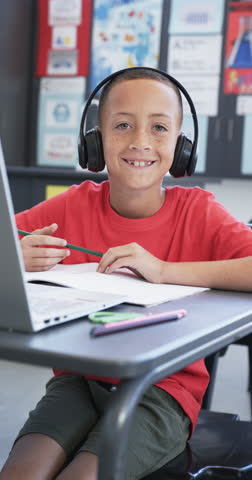 Vertical video: In school, child wearing headphones using laptop and writing in classroom. Education, technology, learning, studying, childhood, slow motion