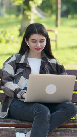 Vertical video of woman in headset typing on the laptop computer on the bench in the park. Female makes conference video call on laptop computer talks with web tutor, online teacher in webcam chat.