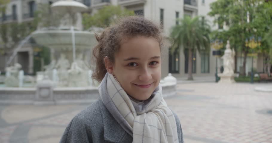 Portrait of a beautiful 12 old girl in a spring gray coat and white scarf,girl teenager gently smiling while looking at camera, standing in a park on an autumn day with an urban fountain in background