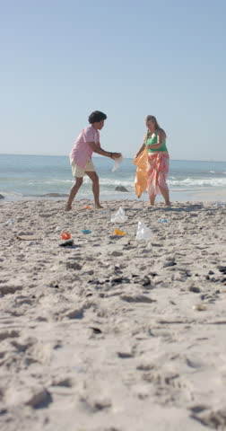 Vertical video: Cleaning beach, group of friends picking up trash from sand near ocean. Environment, volunteering, teamwork, beach cleanup, sustainability, community, slow motion