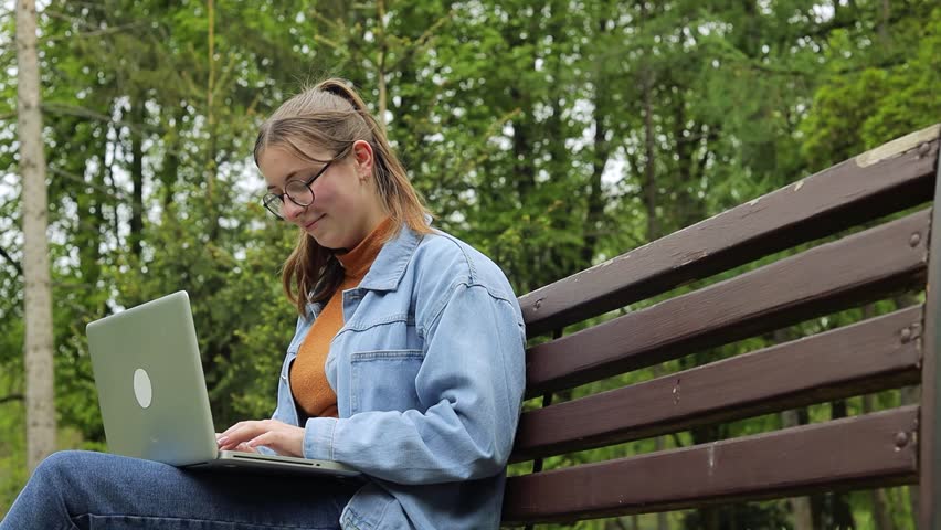 Young female student working remotely on laptop while sitting on wooden park bench, surrounded by lush green trees and enjoying peaceful outdoor environment


