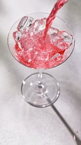 fresh red drink pouring into a cocktail glass with ice cubes on a gray sunny restaurant tabletop, slow motion
