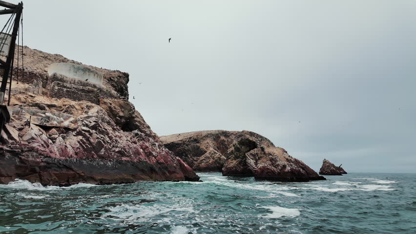 Dramatic views of the Ballestas Islands in Peru, with rugged cliffs and flocks of birds soaring under a cloudy Pacific sky.